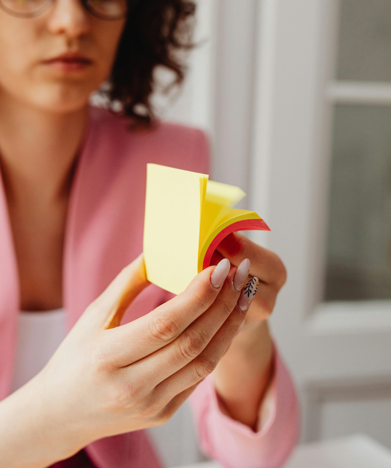 professional woman with brown hair wearing glasses holding a stack of yellow and pink post it notes in her hands whilst flicking through them professional woman with brown hair wearing glasses holding a stack of yellow and pink post it notes in her hands whilst flicking through them