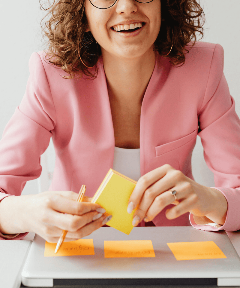 Brown haired woman, smiling, holding a stack of yellow post it notes above a folded laptop with three strategy notes already stuck to it. Brown haired woman, smiling, holding a stack of yellow post it notes above a folded laptop with three strategy notes already stuck to it.